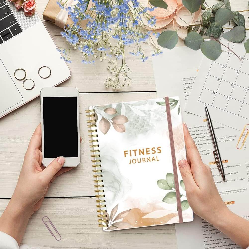 Person holding Herbionyx Fitness Journal at a desk with a laptop and phone, surrounded by stationery and flowers.